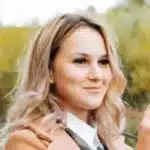 Eine Frau Mit Gewelltem Blondem Haar Und Einem Kamelfarbenen Mantel L&auml;chelt, W&auml;hrend Sie Eine Kamera Im Freien H&auml;lt Und Momente Nach Beendigung Ihrer Reinigungsarbeiten Festh&auml;lt, Wobei Die Gr&uuml;nen B&auml;ume Im Hintergrund Verschwimmen.