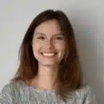 Eine Junge Frau Mit Glattem Braunem Haar Und Grauem Pullover Steht Mit Einem Strahlenden L&auml;cheln Vor Einem Schlichten Hellen Hintergrund Und Ist Bereit, Erstklassige Reinigungsdienste Anzubieten.