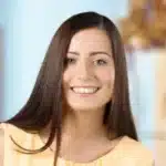 Eine Junge Frau Mit Langen Braunen Haaren Und Einem Strahlenden L&auml;cheln Tr&auml;gt Ein &Auml;rmelloses Gelbes Oberteil. Sie Steht In Einem Sanft Beleuchteten Innenraum Mit Unscharfem Hintergrund Und Verk&ouml;rpert Perfekt Das Einladende Gesicht Eines Vertrauensw&uuml;rdigen Reinigungsdienstes.