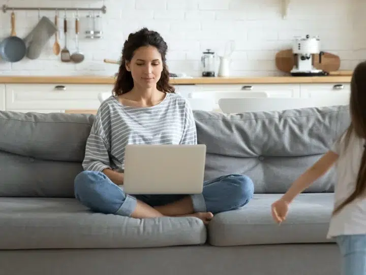 Eine Frau Sitzt Im Schneidersitz Auf Einem Grauen Sofa Und Benutzt Einen Laptop In Einer Hellen Küche, Während Sich Ein Kleines Kind In Der Nähe Bewegt, Das Teilweise Im Vordergrund Zu Sehen Ist.