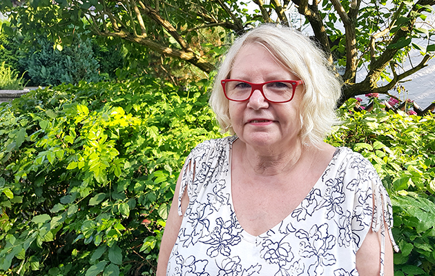 Ältere Frau mit roten Brillen vor grünem Busch, sonniger Tag, Thema: Reinigungskraft.