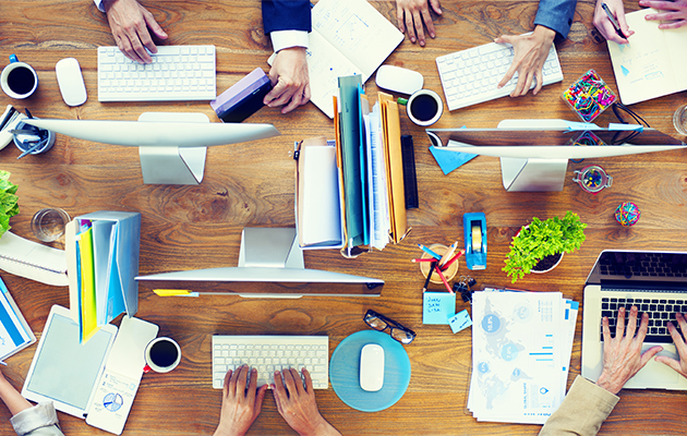 Bürotisch mit Computern, Dokumenten, Pflanzen und Händen. Thema: Reinigungskraft im Büro.