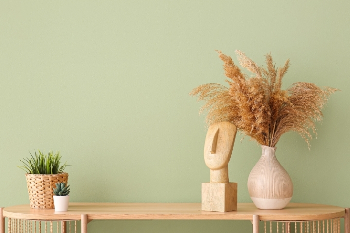 Holztisch mit Dekoration: Vase mit Trockenblumen, Pflanze, Skulptur, vor grünem Hintergrund.