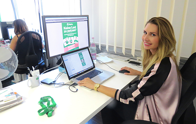 Frau am Schreibtisch mit Laptop, Monitor zeigt "Keine Lust zu putzen?" Werbung.