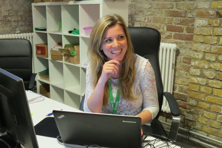 Frau am Schreibtisch mit Laptop, lächelnd, vor Regal. Thema: Büro, Reinigungskraft.