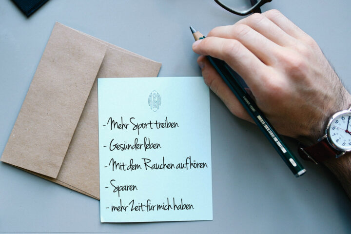 Hand schreibt Vorsätze auf Notizblock; Brille und Umschläge daneben. Thema: reinigung.