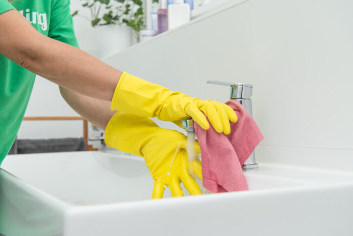Gelbe Handschuhe reinigen Wasserhahn mit rosa Tuch, reinigungskraft in grünem Shirt.