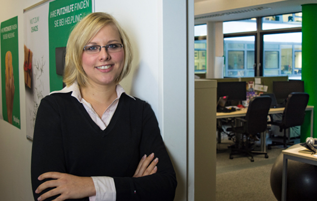 Frau mit Brille lächelt im Büro neben Plakat über Putzfrau-Dienste.