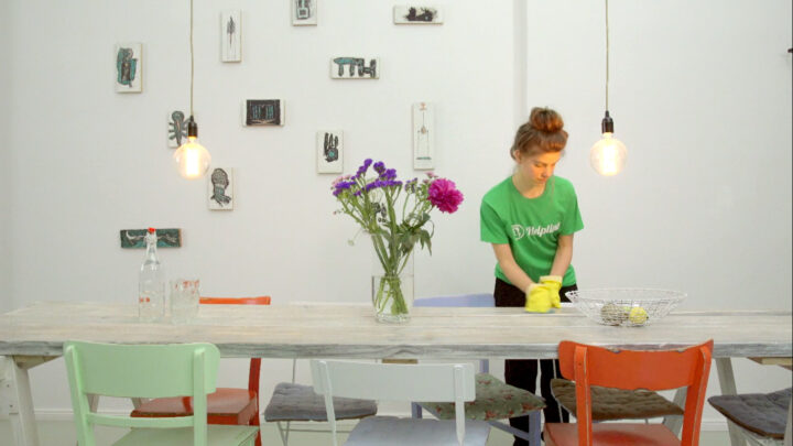 Putzfrau reinigt Tisch in modernem Raum mit Blumen, Wandkunst und Hängelampen.