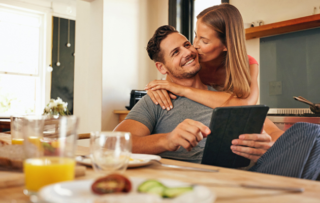 Paar in Küche, Frau umarmt Mann mit Tablet, Frühstück auf Tisch, reinigung im Hintergrund.
