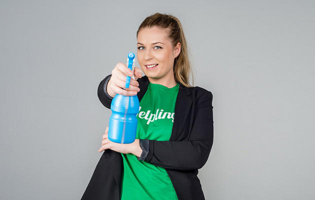 Frau in grüner Helpling-Shirt hält blauen Reiniger, lächelnd vor grauem Hintergrund.