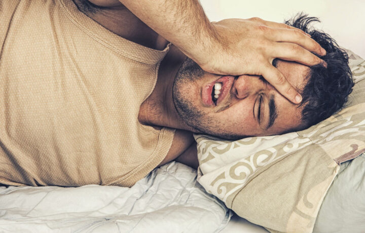 Mann im Bett hält sich den Kopf, wirkt müde oder gestresst. Kissen im Hintergrund.
