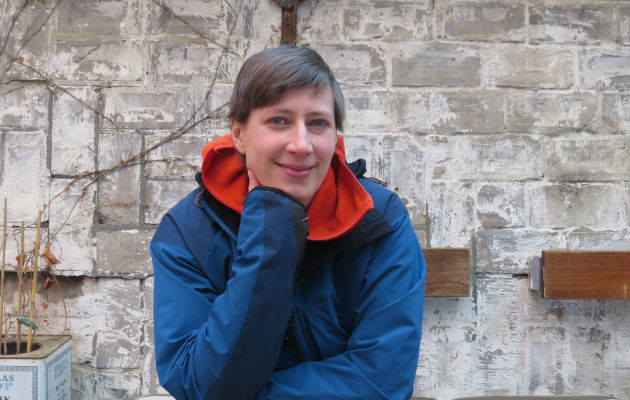 Person in blauer Jacke vor grauer Ziegelwand, lächelnd. Thema: Reinigungskraft.