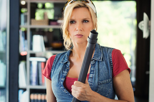 Frau in Jeansweste hält Staubsaugerrohr, Bücherregal im Hintergrund, reinigungskraft.