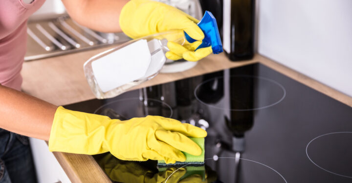Person mit gelben Handschuhen reinigt Herd mit Sprühflasche und Schwamm.