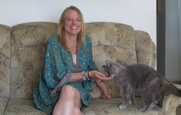 Frau in blauem Kleid streichelt graue Katze auf Sofa, lächelnd. Thema: Putzfrau.