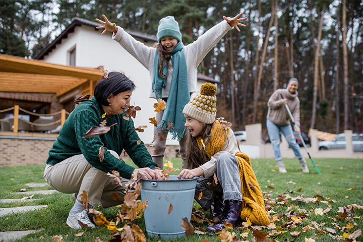 Familie Sammelt Herbstblätter Im Garten, Während Eine Person Im Hintergrund Mit Einem Rechen Putzt.