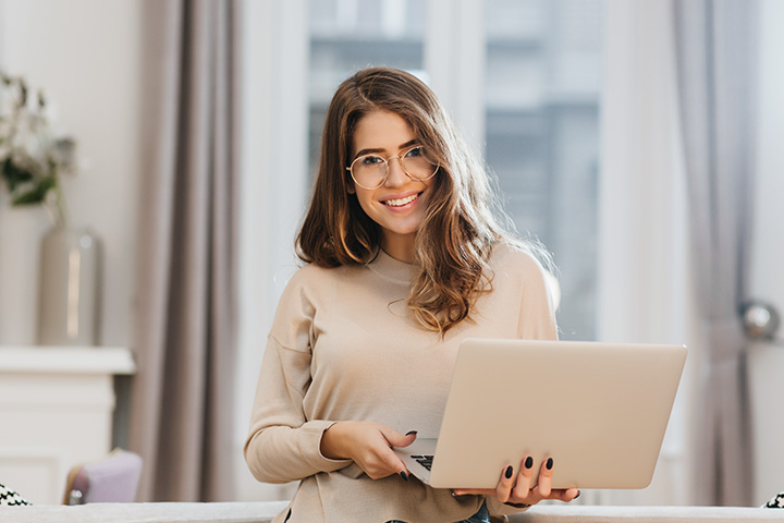 Frau mit Laptop lächelt, sitzt auf Sofa. Moderne Wohnung, Vorhänge, Vase im Hintergrund.