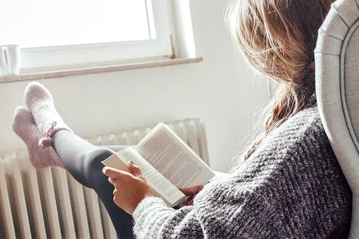 Frau liest entspannt ein Buch am Fenster, trägt Socken. Reinigen nicht erforderlich.