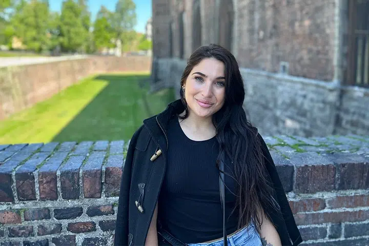 Frau steht vor Ziegelmauer, lächelt, trägt schwarzen Mantel. Hintergrund: Grünfläche.