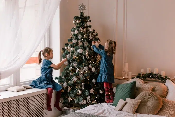 Kinder schmücken Weihnachtsbaum im Wohnzimmer, dekoriert mit Lichtern und Kugeln.