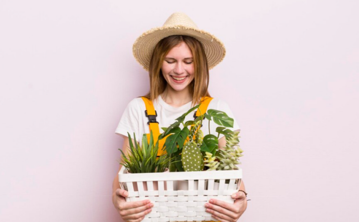 Frau mit Strohhut hält einen Korb voller Pflanzen vor rosa Hintergrund, lächelnd. Reinigungskraft.