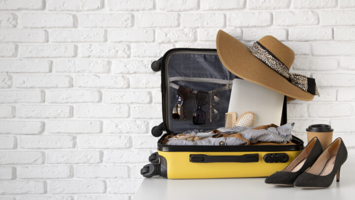 Gelber Koffer mit Sonnenhut, Sonnenbrillen, Laptop und Schuhen vor weißer Ziegelwand. Reinigung.