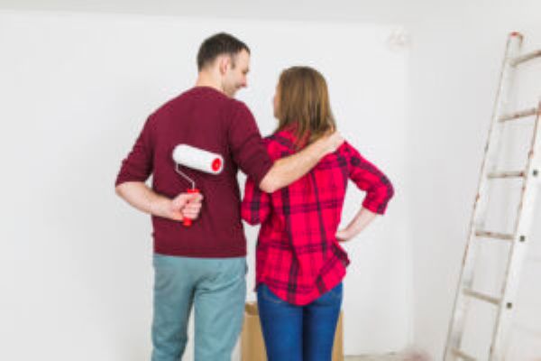 Paar mit Farbroller vor weißer Wand, Leiter rechts. Renovierung und putzen geplant.