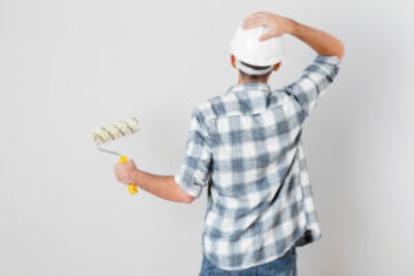 Mann mit Karohemd und Helm hält Farbroller, steht vor weißer Wand, Hand am Kopf.