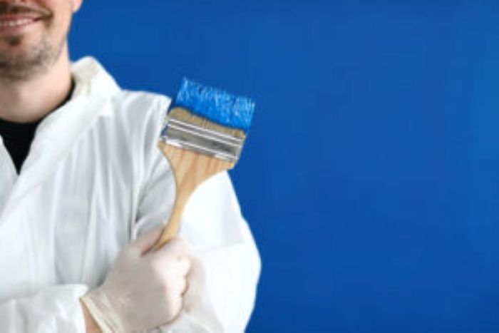 Mann in Schutzanzug hält Pinsel vor blauer Wand, ideal für Reinigungskraft-Themen.