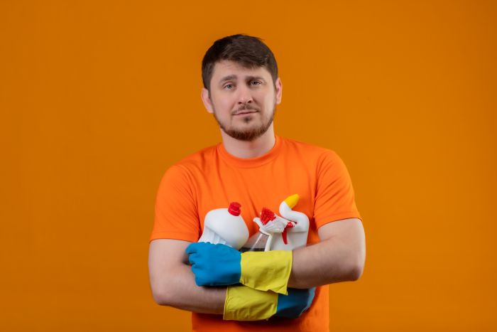 Young Man Wearing Orange T Shirt Rubber Gloves Holding Cleaning Supplies With Serious Confident