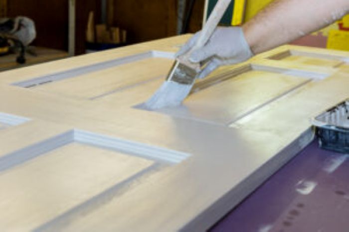 Hand mit Pinsel streicht weiße Farbe auf Holztür, Vorbereitung für Reinigung.