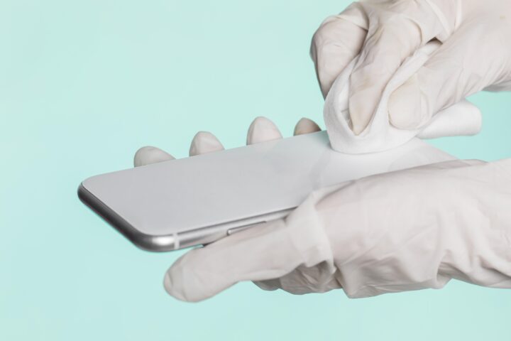 Hand mit Handschuhen reinigt Smartphone mit Tuch, Fokus auf Sauberkeit und Hygiene.