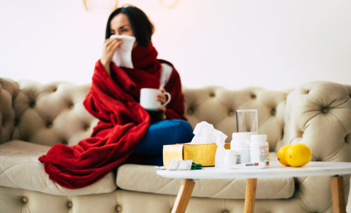 Frau mit roter Decke auf Sofa, Taschentücher und Medikamente auf Tisch, reinigungskraft.