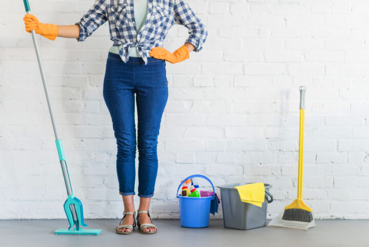 Putzfrau mit Wischmopp, Eimer und Besen vor weißer Wand, bereit zum Reinigen.