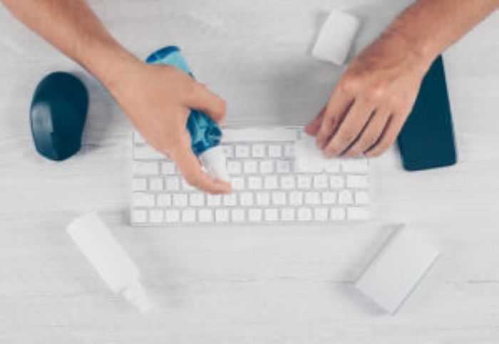 Tastaturreinigung mit Spray und Tuch, Hände putzen Schreibtisch, Smartphone und Maus daneben.