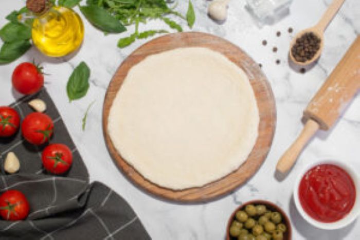 Pizzateig auf Holzbrett, umgeben von Tomaten, Oliven, Basilikum, reinigung, Nudelholz.