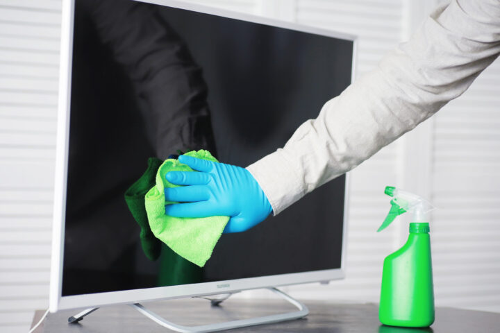 Hand mit Handschuh reinigt Bildschirm mit grünem Tuch, Sprühflasche daneben, Reinigung.