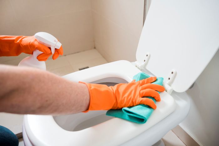 Reinigungskraft mit Handschuhen putzt Toilette mit Spray und Tuch.