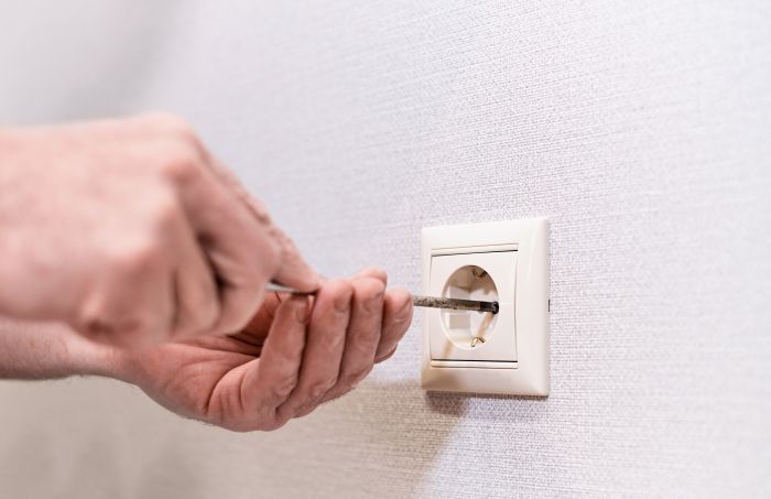 Hand repariert Steckdose mit Schraubenzieher an weißer Wand. Reinigungskraft nicht erkennbar.
