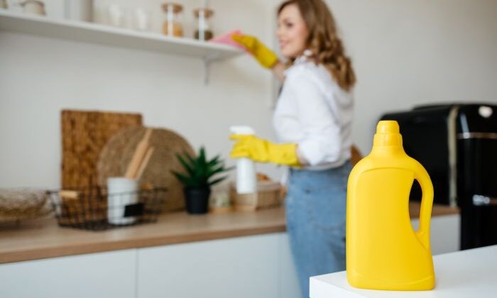 Gelbe Reinigungsflasche Im Vordergrund, Putzfrau Reinigt Regal Im Hintergrund.