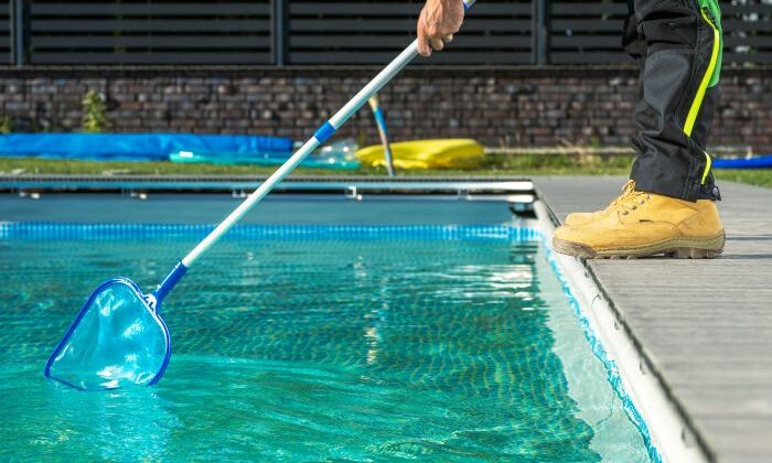 Person reinigt Pool mit Kescher, trägt gelbe Stiefel und schwarze Hose.
