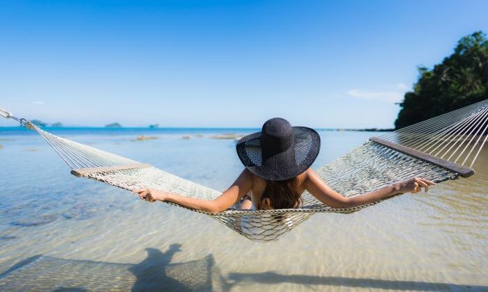 Person in Hängematte über Wasser, trägt schwarzen Hut, entspannter Strandtag.