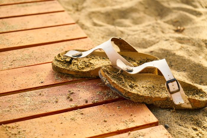 Sandalen im Sand neben Holzplanken, symbolisiert Reinigung nach Strandbesuch.