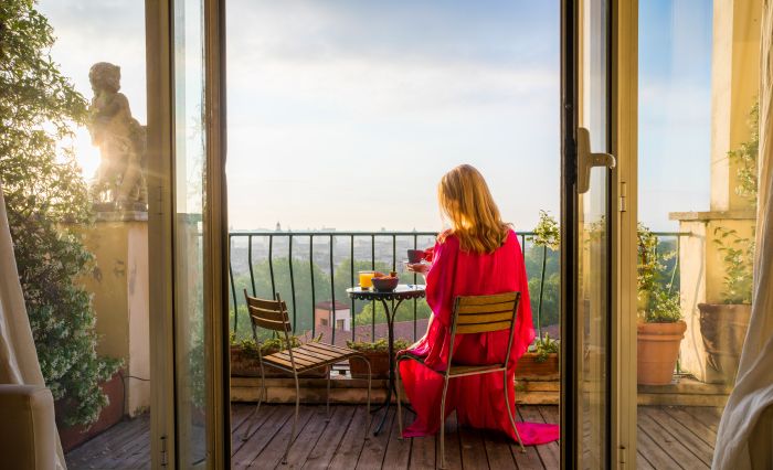 Frau in rotem Kleid frühstückt auf Balkon mit Aussicht, umgeben von Pflanzen.
