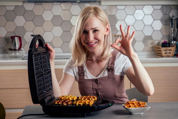 Frau lächelt, zeigt OK-Zeichen, Waffeln im Waffeleisen, Küche, putzen.