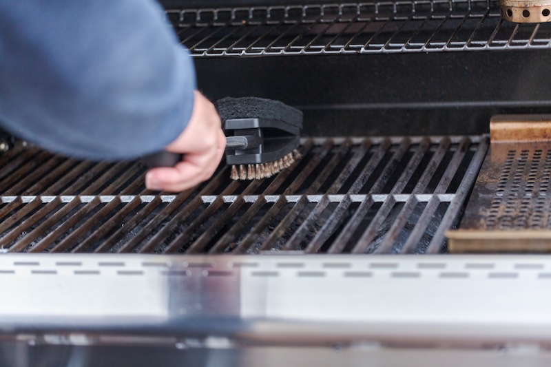 A Hand Cleans A Grill Grate With A Metal Brush. The CloseUp Shows Shiny Metallic Bars And The Brush's Bristles, Suggesting Care And Maintenance.