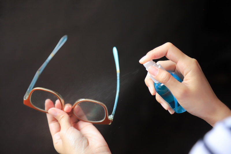 Hände Halten In Der Einen Hand Eine Brille Und Sprühen In Der Anderen Linsenreiniger Aus Einer Blauen Flasche Vor Dunklem Hintergrund.