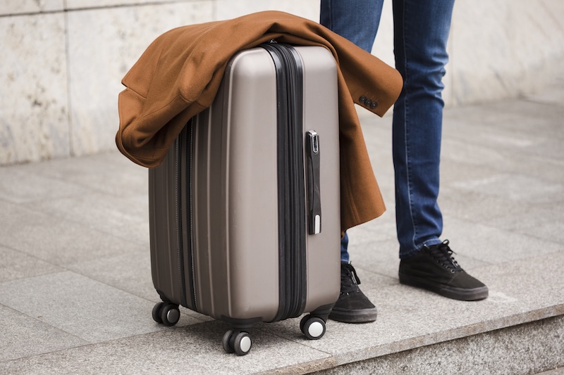 A Silver Suitcase With Four Wheels Stands On A Stone Pavement. A Brown Coat Is Draped Over It. A Person Wearing Jeans And Black Shoes Is Next To The Suitcase.
