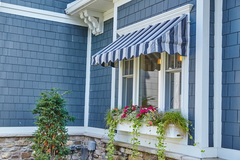 Charmante Blaue Häusercke Mit Gestreifter Markise, Fensterkasten Voller Rosa, Roter Blumen Und Üppigem Grün Ruft Eine Gemütliche, Einladende Atmosphäre Hervor.